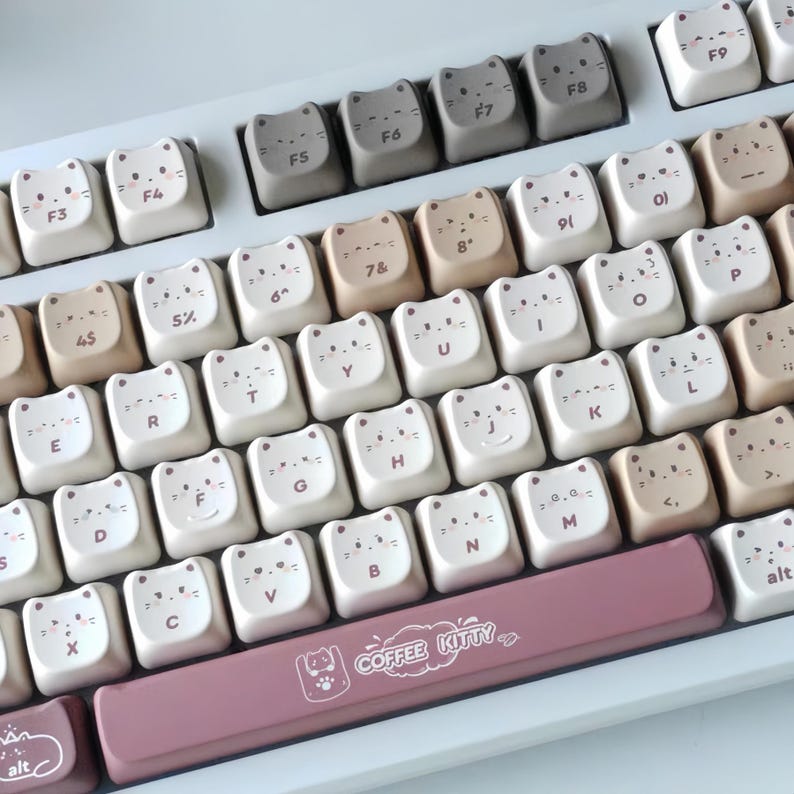 Puede incluir: Un teclado con teclas en forma de gato en tonos blanco, beige y gris. Las teclas presentan caras de gato y letras. La barra espaciadora es de color rosa empolvado y tiene el texto "COFFEE KITTY".