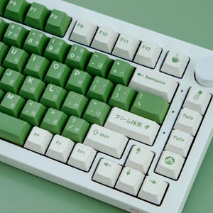 May include: A white mechanical keyboard with green and white keycaps. The keycaps have Japanese characters and English letters. The keyboard has a green theme, with green keys and a white frame. The keyboard is on a green surface.