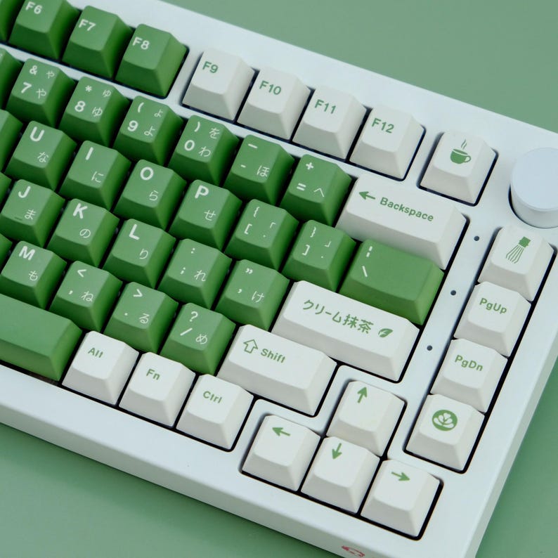 May include: A white mechanical keyboard with green and white keycaps. The keycaps have Japanese characters and English letters. The keyboard has a green theme, with green keys and a white frame. The keyboard is on a green surface.