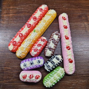 May include: A selection of miniature eclairs in various colours, including red, yellow, orange, pink, brown, purple, black, and green. Each eclair is decorated with white icing and fruit toppings, arranged on a dark wooden surface.