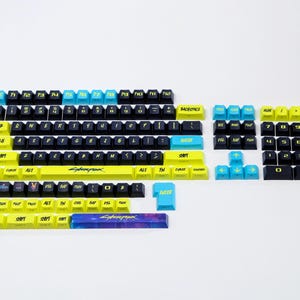 May include: A set of Cyberpunk-themed keyboard keycaps in various sizes and layouts. The keycaps are black, yellow, and blue, with the word "Cyberpunk" printed on the spacebar. The keycaps are designed for mechanical keyboards.