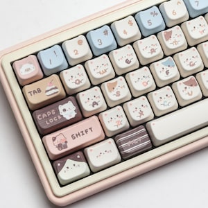 May include: A pastel pink and cream-colored keyboard with custom keycaps. The keycaps feature cat faces and other cute designs. The keyboard has a standard layout with number keys, letters, and function keys.