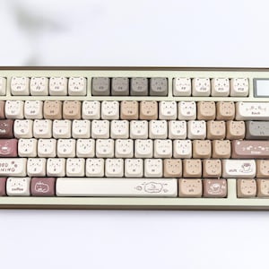 Puede incluir: Un teclado mec&aacute;nico beige y marr&oacute;n con teclas personalizadas. Las teclas presentan caras de oso e ilustraciones con tem&aacute;tica de caf&eacute;. El teclado tiene forma rectangular con esquinas redondeadas y el texto "Good Morning".