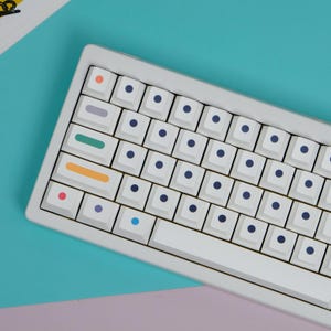 May include: A white mechanical keyboard with light blue, orange, green, and purple accent keys. The keyboard has white keycaps with dark blue dots. The keyboard is on a teal and purple background.