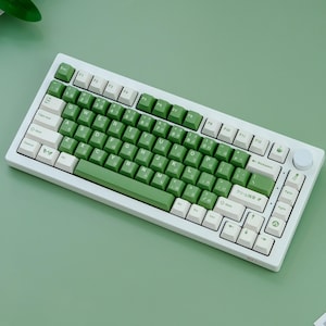May include: A white mechanical keyboard with green and white keycaps. The keyboard has a compact layout with Japanese characters on the keys. The keycaps are a mix of white and green, creating a visually appealing contrast. The keyboard is on a green surface.