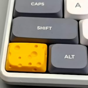Könnte beinhalten: Nahaufnahme einer Computertastatur mit grauen und weißen Tasten. Die Tasten "SHIFT", "ALT" und "CAPS" sind grau. Eine gelbe Taste in Form von Schweizer Käse befindet sich in der Mitte. Die Tasten befinden sich in einem weißen Rahmen.
