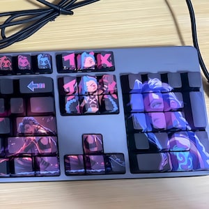 May include: A custom mechanical keyboard with keycaps featuring characters from the animated series Arcane. The keycaps display images of characters in shades of purple, blue, and pink. The keyboard has a gray base and a black cable.