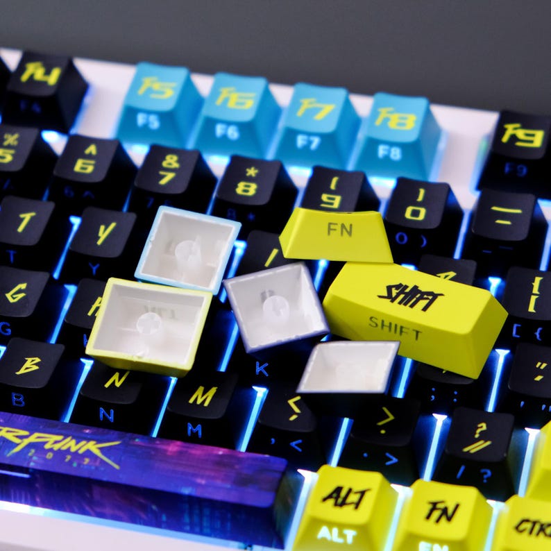 May include: Close-up of a mechanical keyboard with various keycaps. The keyboard features black, blue, and yellow keycaps, with some keys removed. The "SHIFT" key is yellow with black text. The keyboard is lit with blue backlighting.
