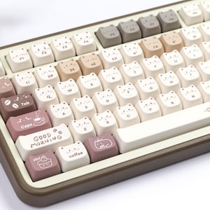 Puede incluir: Un teclado mec&aacute;nico con teclas en forma de gato en tonos blancos, beige y marr&oacute;n. Algunas teclas presentan caras de gato, tazas de caf&eacute; y la frase &laquo;GOOD MORNING&raquo;. El teclado tiene un marco marr&oacute;n y una barra espaciadora blanca.