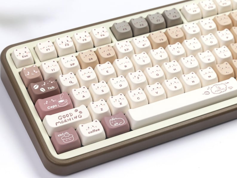 Puede incluir: Un teclado mec&aacute;nico con teclas en forma de gato en tonos blancos, beige y marr&oacute;n. Algunas teclas presentan caras de gato, tazas de caf&eacute; y la frase &laquo;GOOD MORNING&raquo;. El teclado tiene un marco marr&oacute;n y una barra espaciadora blanca.