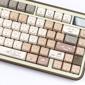 Puede incluir: Un teclado mec&aacute;nico beige y marr&oacute;n con teclas en forma de gato. Las teclas son de varios tonos de beige y marr&oacute;n, con caras de gato en cada tecla. El teclado tiene una peque&ntilde;a pantalla y la palabra "Enter" en una tecla.