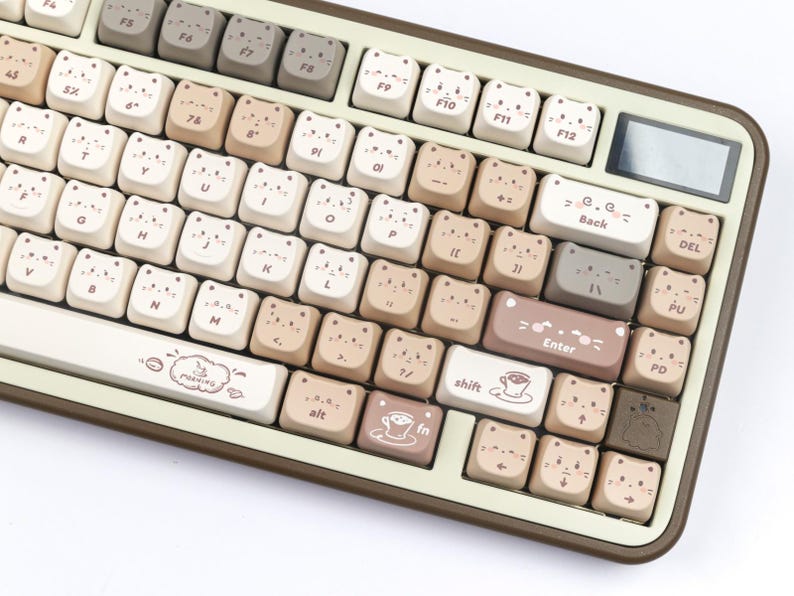 Puede incluir: Un teclado mec&aacute;nico beige y marr&oacute;n con teclas en forma de gato. Las teclas son de varios tonos de beige y marr&oacute;n, con caras de gato en cada tecla. El teclado tiene una peque&ntilde;a pantalla y la palabra "Enter" en una tecla.