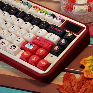 May include: Custom mechanical keyboard with a red and white frame and colorful keycaps. The keycaps feature various designs, including Pokeball and Pokemon character graphics. Some keys have Japanese characters. The keyboard is on a brown surface.