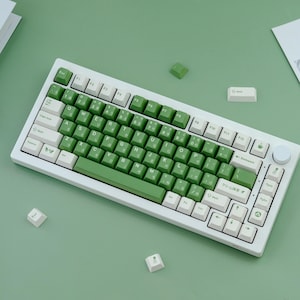 May include: A white mechanical keyboard with green and white keycaps. The keyboard has a green accent bar and a rotary dial. Several loose keycaps are scattered around the keyboard on a green surface.