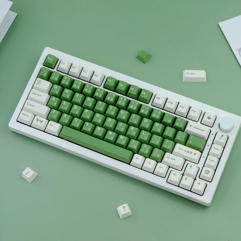 May include: A white mechanical keyboard with green and white keycaps. The keyboard has a green accent bar and a rotary dial. Several loose keycaps are scattered around the keyboard on a green surface.