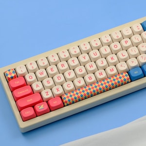 May include: A beige mechanical keyboard with white keycaps and pink, blue, and patterned accent keys. The keyboard has a standard QWERTY layout with numbers and symbols. The keycaps have pink lettering. The keyboard is on a light blue surface.