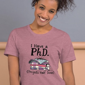 Könnte beinhalten: Ein heidefarbenes T-Shirt mit einer humorvollen Grafik einer Person, die auf einem Stapel Bücher schläft. Der Text auf dem Shirt lautet "I Have a PhD. (Projects Half Done)"
