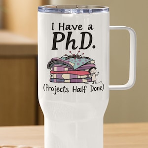 Könnte beinhalten: Weiße Reisebecher mit einem schwarz-weißen Design, auf dem "Ich habe einen Doktortitel. (Projekte halb fertig)" steht. Das Design zeigt einen Stapel Stoffquadrate mit einer Nadel und einem Faden, die aus der Oberseite herausragen. Eine kleine Person steht auf dem Stoffquadraten und hält eine Nadel und einen Faden.