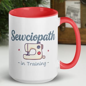 Puede incluir: Taza de cerámica blanca con borde y asa rojos. La taza tiene un gráfico de una máquina de coser y el texto "Sewciopath - in Training -" impreso en ella.