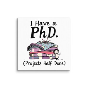 May include: A white canvas print with black text that reads "I Have a PhD. (Projects Half Done)". A cartoon illustration of a pile of fabric with pins and a person standing next to it.