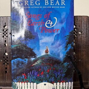 May include: A hardcover book titled "Songs of Earth and Power" by Greg Bear. The cover features a painting of a person on horseback atop a hill with a white picket fence and colorful flowers. The background is a blue sky with mountains.