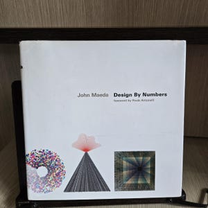 Op de afbeelding: Een wit boek getiteld "Design By Numbers" van John Maeda, met een voorwoord van Paola Antonelli. De omslag toont abstracte geometrische ontwerpen in verschillende kleuren, waaronder een donutvormig patroon, een piramide en een vierkant.