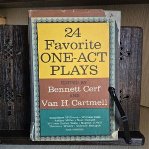 Może przedstawiać: Zabytkowa książka zatytułowana "24 Favorite One-Act Plays" zredagowana przez Bennetta Cerfa i Vana H. Cartmella. Książka ma żółtą, brązową i zieloną okładkę z nazwiskami dramaturgów. Książka jest podparta drewnianym stojakiem na książki.