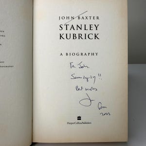 May include: An open book displaying the title "STANLEY KUBRICK A BIOGRAPHY" by John Baxter. The book is signed "To John, Serendipity!! Best wishes" with a signature and the year 2022. The publisher's logo is at the bottom.