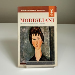 May include: A vintage art book titled "Modigliani" featuring a portrait on the cover. The book has a brown and cream cover with the title in large red letters. The cover also includes the text "A Mentor-UNESCO Art Book".