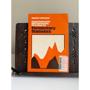 May include: An orange book titled "Study Guide with Self-Correcting Exercises for Elementary Statistics" by Robert Johnson. The book has a white and orange graphic on the cover and is displayed in a dark wooden book holder.