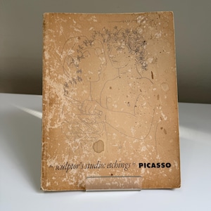 May include: A vintage book cover with a sketch of two figures, one holding a knife. The cover is tan with the title "the sculptor's studio: etchings by PICASSO" in black text. The cover has a worn, aged appearance.