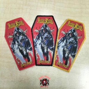 Puede incluir: Tres parches bordados con forma de ataúd que presentan el logotipo de Judas Priest y el texto Hero, Hero. Cada parche muestra una figura central en tonos negros y grises sobre un fondo rojo. Los bordes son de color rojo, negro y dorado.