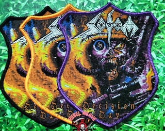 May include: A shield-shaped embroidered patch featuring the Sodom band logo and the text Decision Day. The design depicts a skeletal figure with a blue eye and a rifle against a yellow and orange background, finished with a purple border.