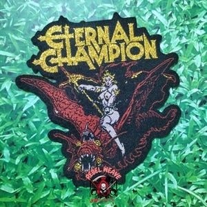 May include: A black embroidered patch featuring the gold text ETERNAL CHAMPION at the top. The design depicts a figure riding a large, red, winged creature with sharp teeth. At the bottom, the text REBEL WEAVE appears above a small skull logo.
