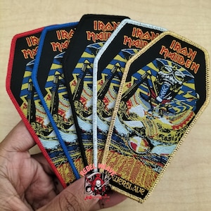 Puede incluir: Un conjunto de cinco parches bordados con forma de ataúd que presentan la ilustración del álbum Powerslave de Iron Maiden con un faraón egipcio. Cada parche tiene un borde distinto en hilo rojo, azul, negro, blanco y dorado.