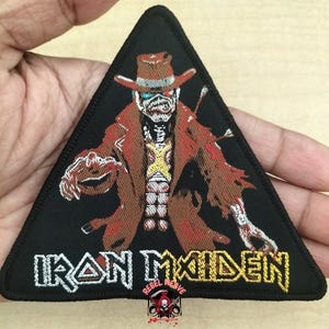 Könnte beinhalten: Ein dreieckiger, schwarzer Aufnäher mit einer gestickten Illustration einer skelettartigen Figur in einem braunen Hut und Mantel. Der Text "IRON MAIDEN" ist in Weiß und Gold gestickt. Kleines Logo mit Totenkopf und gekreuzten Knochen unten.