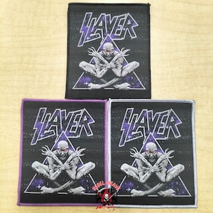 May include: Three rectangular fabric patches featuring the Slayer logo in purple and white text above a graphic of a seated figure with crossed arms inside a purple triangle. The background is black with a starry space pattern and white borders.