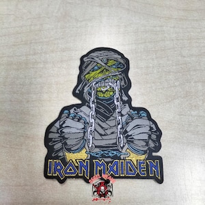 May include: A rectangular embroidered patch featuring a mummy-like figure with green skin, wrapped in grey bandages, and wearing silver chains. The text IRON MAIDEN is embroidered in blue with a gold outline at the bottom of the patch.