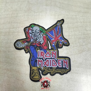 May include: An embroidered patch featuring a skeletal figure in a red jacket and blue trousers holding a tattered Union Jack flag. The text Iron Maiden and The Trooper is displayed in a bold, stylized font with a gold border on a black background.