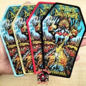 Könnte beinhalten: Vier Sarg-förmige Aufnäher mit dem Text "The Black Dahlia Murder" und einem grafischen Design. Die Aufnäher haben unterschiedlich farbige Ränder: hellblau, gold, rot und schwarz. Die Aufnäher werden in einer Hand gehalten.