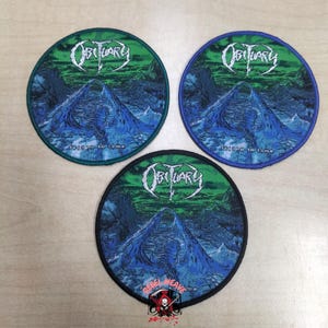 May include: Three circular embroidered patches featuring the Obituary logo in white text above a blue mountain landscape and green sky. Each patch displays the text Frozen in Time. The patches have green, blue, and black stitched borders respectively.