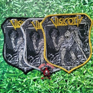 May include: Three shield-shaped embroidered patches featuring the text Visigoth in yellow. The design depicts a skeletal figure in armour holding a sword and a flame, surrounded by skulls. The patches have black, silver, and yellow thread details.