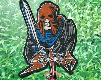 May include: An embroidered patch featuring a hooded figure in a brown mask and black cloak holding a silver sword. The word SODOM is stitched in a stylized orange and white font at the bottom. The design includes blue accents on the clothing.