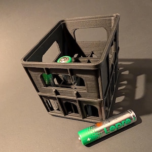 May include: A black plastic crate with a grid pattern, designed to hold small items. The crate is filled with several green and black batteries. A single green and black battery is lying on the surface next to the crate.