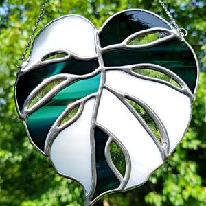 Peut inclure: Feuille en vitrail en forme de cœur, composée de verre blanc et vert. La feuille est encadrée de métal argenté et suspendue à une chaîne. Le design rappelle une feuille de plante Monstera.