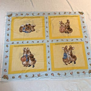 May include: A light blue fabric panel featuring four yellow-bordered squares with Peter Rabbit illustrations. Text includes: Peter and Benjamin Bunny watching a duckling, Peter in the red spotted handkerchief with Benjamin Bunny, and Benjamin Bunny giving Peter Rabbit a flower.
