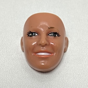 May include: A plastic doll head with a tan complexion. The face has painted features including black eyelashes, blue eyes, and a smiling mouth with pink lips. The head is bald and has a smooth, shiny surface.
