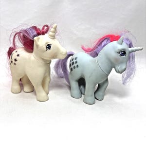May include: Two plastic unicorn figurines with rooted hair. The left unicorn is cream-colored with a star and moon symbol and pink and purple hair. The right unicorn is light blue with a diamond symbol and purple and pink hair.
