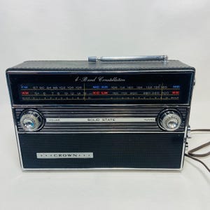 May include: Vintage black and silver Crown 4-Band Constellation radio. The radio features a tuning dial, volume control, and the text "SOLID STATE". The radio has a telescoping antenna and the text "CROWN" on the front.