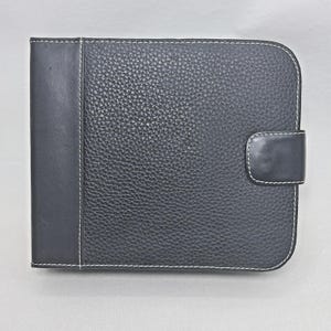 May include: A black leather wallet featuring a textured pebbled finish on the main body and a smooth leather spine. The wallet is accented with white contrast stitching along the edges and includes a matching black leather tab closure.